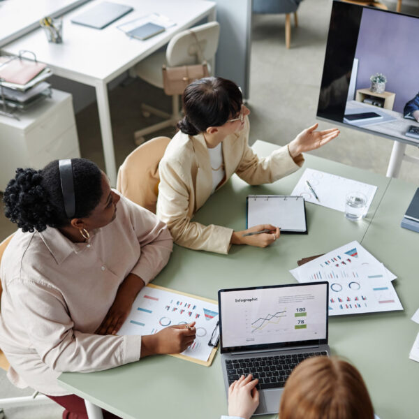 Top down view of diverse business ladies sitting at office desk while discussing annual report during online video call meeting