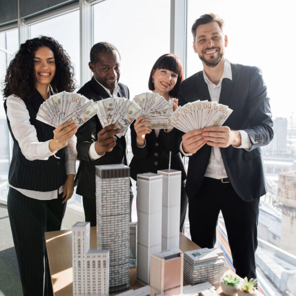 Team of diverse multiethnic business people holding bundle of american dollar cash money, staying near table with 3d skyscraper model at office interior with panoramic city view.